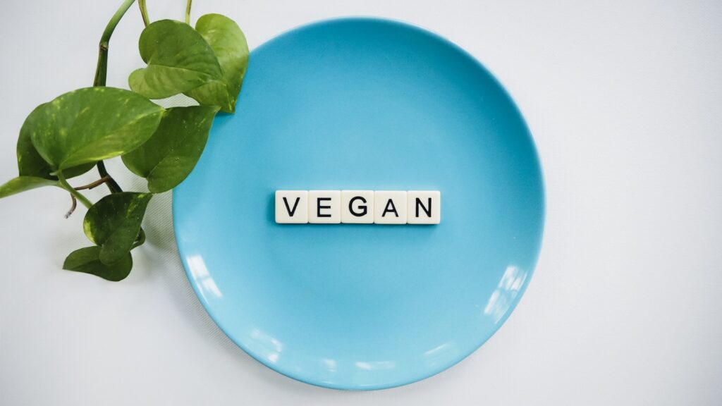 vegan spelled out in scrabble tiles on a blue dinner plate. pothos plant leaves in upper left corner.