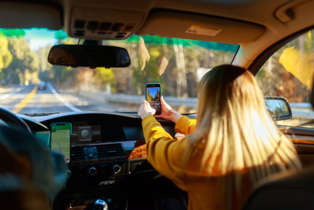 Girl in passenger seat of car holding phone to take picture of road in front of them. Driver seat is filled by someone with their phone propped up on the dash showing a map with directions. It seems like they are on a first road trip.