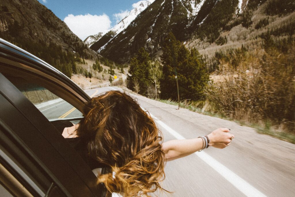 First road trip of a girl, hanging her head out the widow of a car with her hair flowing with the wind. A mountain is in the background, with trees surrounding it.