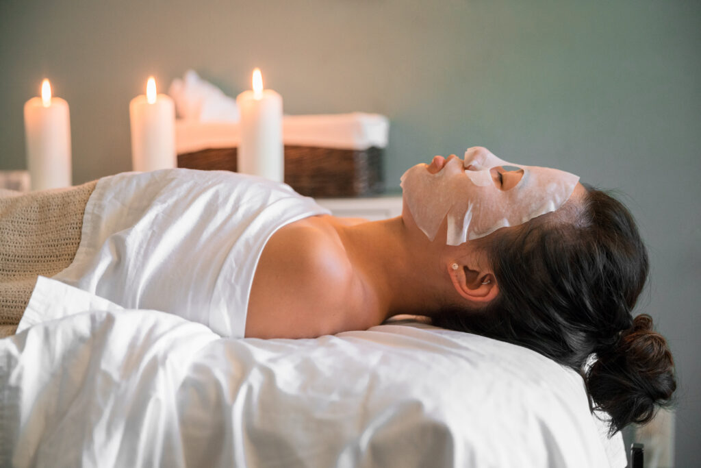 Woman laying on a massage bed, tucked in, with sheet mask on face.