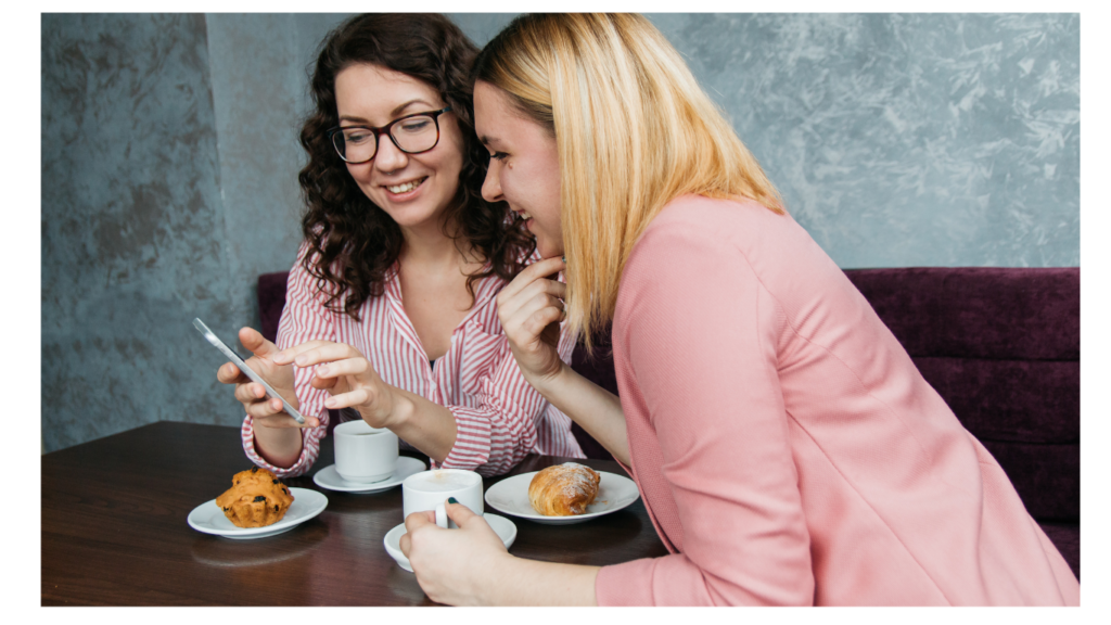 Two women looking at a phone having coffee and muffins