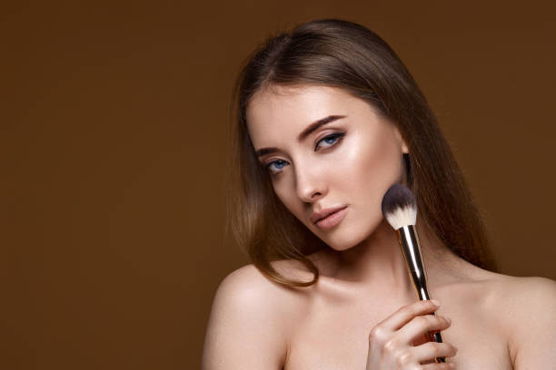 young woman applying highlighter on cheeks with make-up brush on beige background