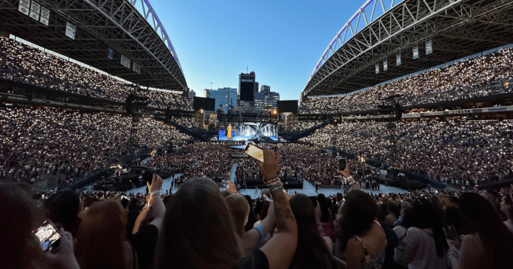 Thousands of people in a stadium, holding their phones up in the air and cheering and listening to live music. The stage is lit up and the Seattle skyline is seen behind the stage. 