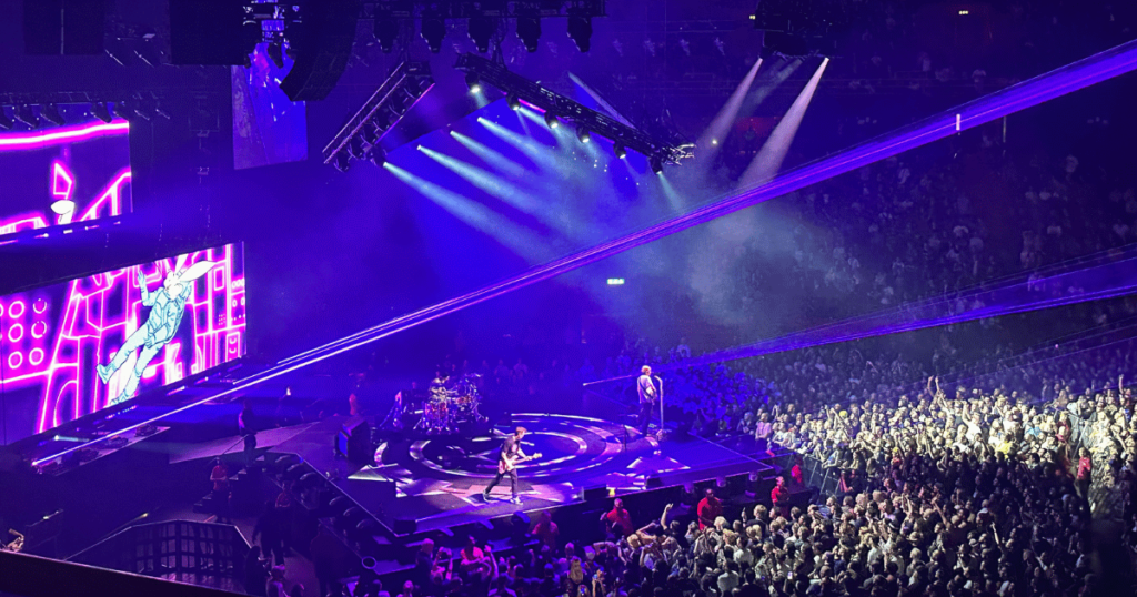 Blue and purple lights emit off the diamond-shaped stage, where one performer stands at the mic, one stands with his guitar on the side, and one sits at the drum set in the middle. Fans are cheering right in front of the stage as they take in the live music.