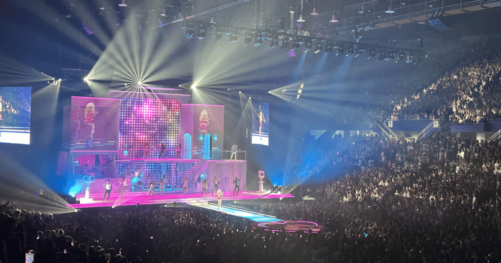 A stage surrounded by fans listening to the music, with the performer at the center in a bright pink, glittery outfit. The stage lights shine everywhere and the blue and pink hues are bright.
