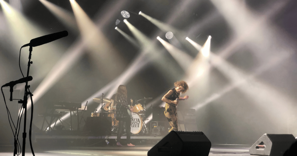 Band on stage with bright stage lights. The lead singer is dancing and the guitarist is barefoot. They are in the middle of a live set.