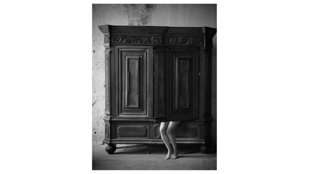 black and white photo of large antique dresser. someone hiding behind the dresser door with only legs showing.