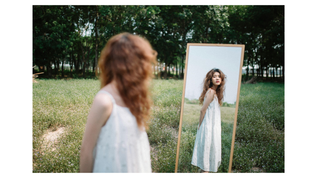 A woman in white dress in a field looking at her reflection in the mirror